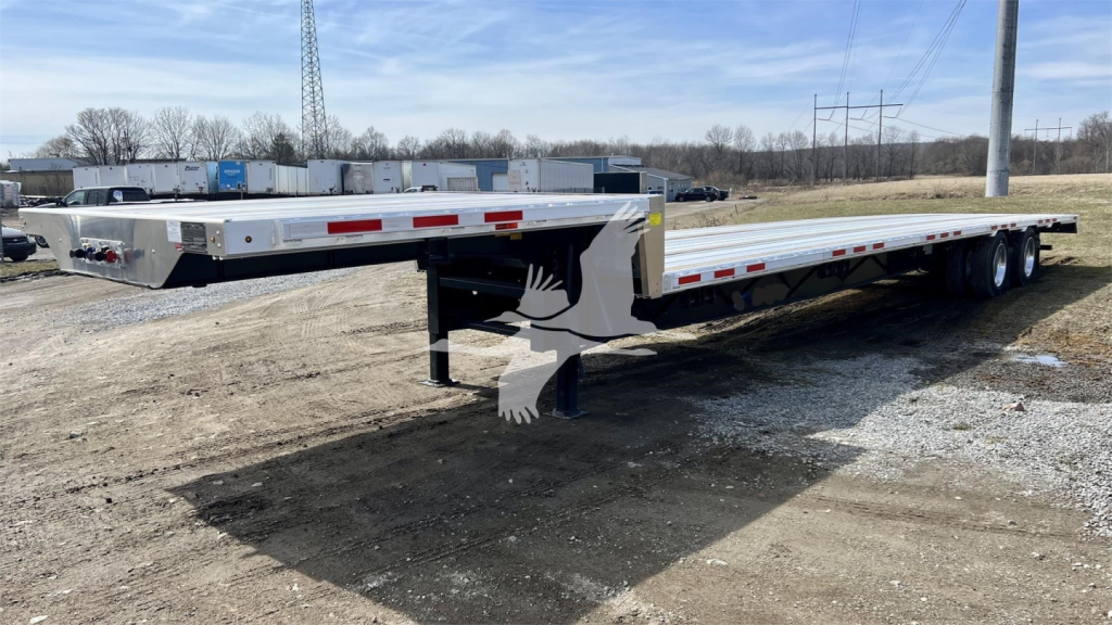 Trailer showing a Transcraft drop deck flatbed with long low-profile deck and triple axle, VIN tag not visible, white and grey color, reflective red/white safety tape along edges.