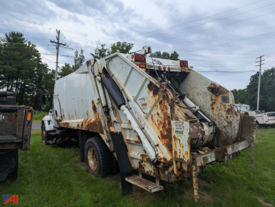 Rear view of a white International WorkStar 7300 20yd trash packer with hydraulic arms and rusted bed, side gate raised, in a gravel lot.