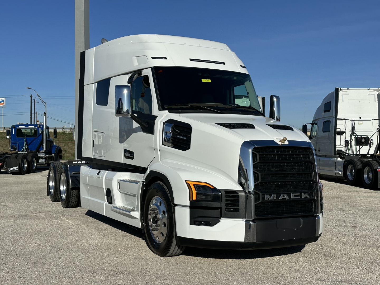 Truck cab with oversized sleeper in white, front grille prominent, Mack badge, daytime; side view showing dual rear axles and chrome mirrors; dealership lot background.