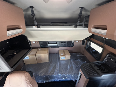 Mack PR64T64 cab interior view with dashboard, overhead bunk area, and steps; several boxes on floor; plastic sheeting covering area; brown and black trim.