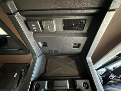 Mack PR64T64 truck interior center console area showing 12V/AC power outlets, USB-style port, and control panel with two round knobs; rubber floor mat visible.