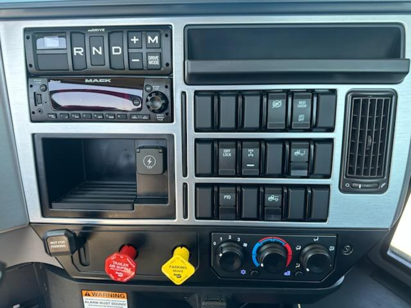 Mack AN64T instrument cluster and control panel showing gear selector R N D with M toggle, radio unit, multiple switch blocks, climate controls, and colored warning knobs.