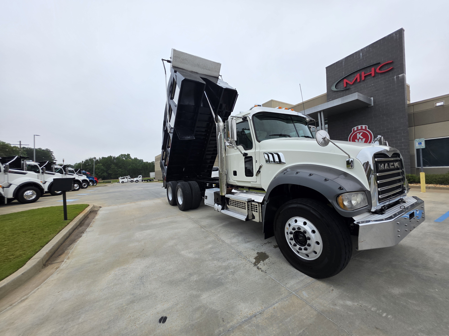 Truck exterior showing a white Mack Granite dump truck with raised dump bed, chrome grille, and dual rear axles. Text on the engine bay area includes Mack logo and model features. Visible signage in background reads MHC and dealership area.