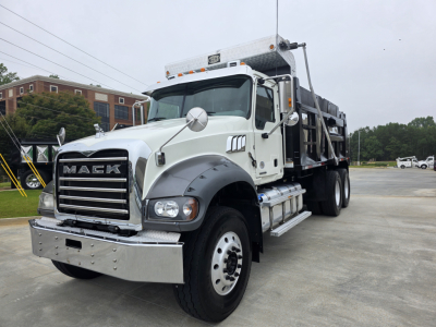 White Mack Granite dump truck in a parking lot; front chrome bumper, grille with MACK letters, round side mirrors, dual fuel tanks under cab, dump bed visible, headlights and warning lights on top.