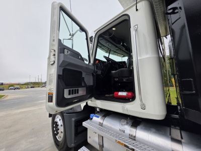 Truck cab interior visible with open door, Mack Granite tractor-truck style. Dashboard and steering wheel seen, passenger seat, red fire extinguisher, metal steps, and fuel tank visible under the door.