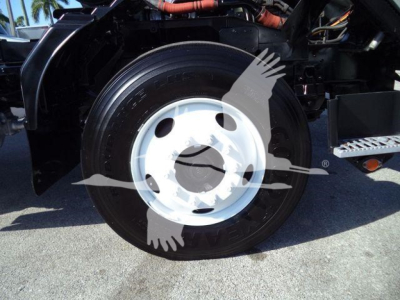 Isuzu truck wheel and front fender view showing black tire with white steel rim; visible engine bay and hydraulic lines in the background.
