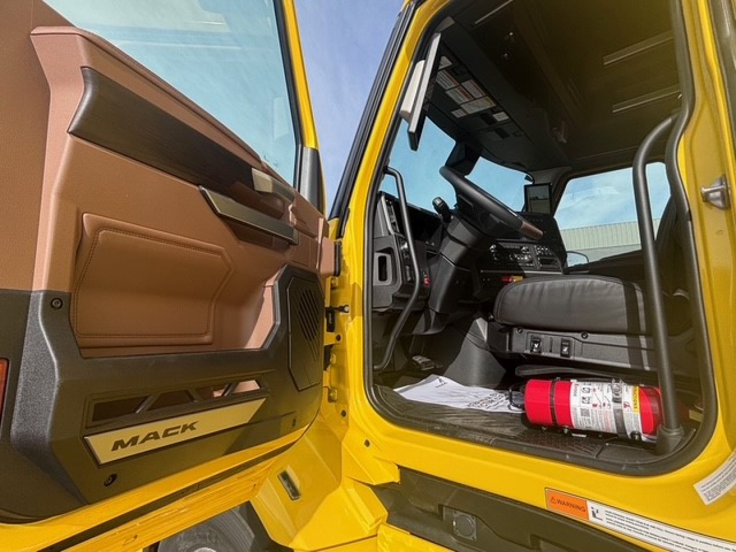 Mack PR64T76H truck cab interior view with open door, yellow exterior, brown interior panels, steering wheel, dashboard, and a fire extinguisher on the floor.