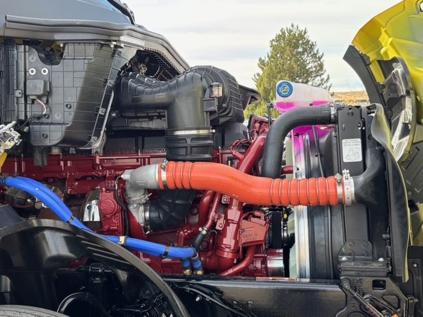 Engine bay of a new Mack PR64T76H truck showing red engine, orange coolant hose, blue hose, radiator, and surrounding components.