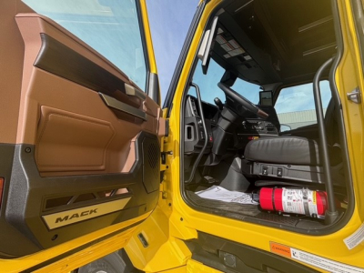 Mack PR64T76H truck cab interior view with open door, yellow exterior, brown interior panels, steering wheel, dashboard, and a fire extinguisher on the floor.