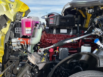 Truck engine bay with exposed red engine block, pink coolant reservoir, hoses, wiring, and yellow exterior; various fluid reservoirs visible.