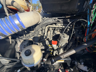 Peterbilt 567 engine bay showing air intake hose, diesel engine, coolant reservoir, and various hoses and wiring. visible components include a round brake reservoir, red cap, and plastic expansion tank.