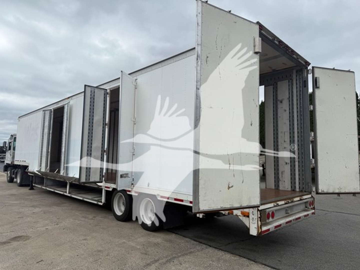 Moving van trailer showing stacked cargo doors open at the rear, white exterior, long rectangular body, tandem rear wheels, and flatbed with rear lights.