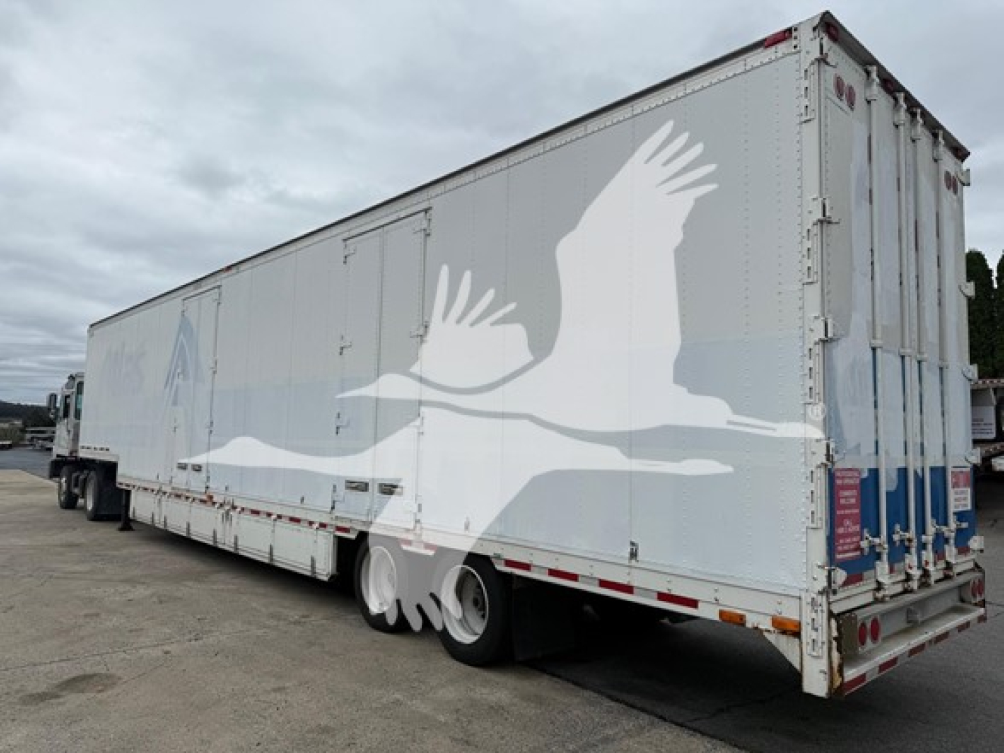 Trailer side view showing large white graphic of flying geese and rear doors. Moving van trailer with dual rear wheels and reflective tape.