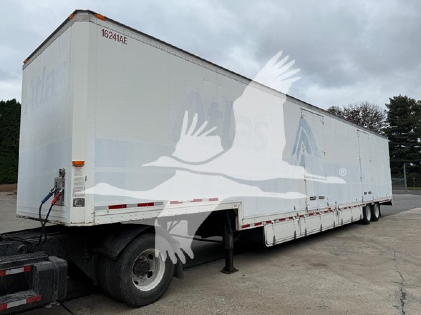 Trailer side view showing moving van with white exterior and subtle blue graphics; rear hitch and landing gear visible; long box trailer with two axles.
