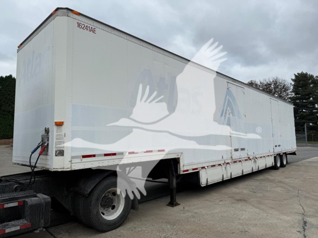 Trailer side view showing moving van with white exterior and subtle blue graphics; rear hitch and landing gear visible; long box trailer with two axles.