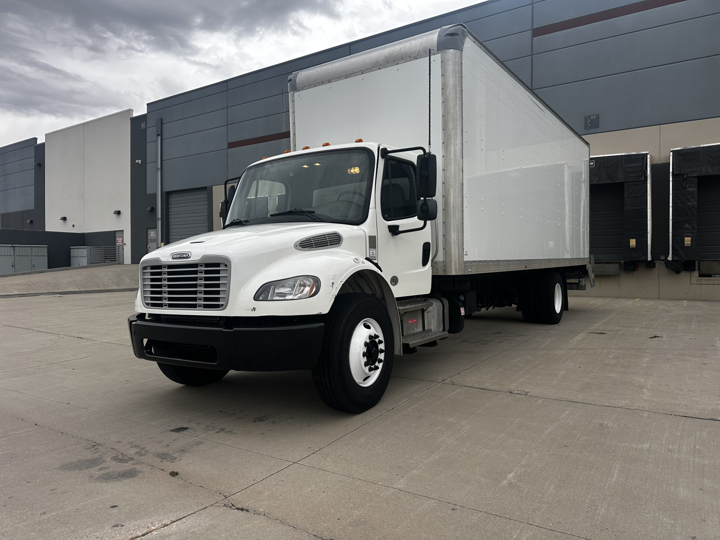 Freightliner M2-106 box truck shown with large white cargo box and cab, front grille, two headlights, and black bumper; dealership lot in background.