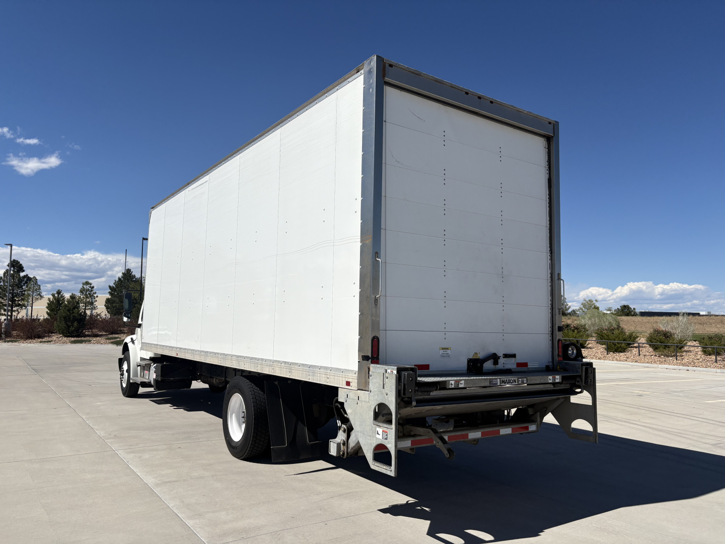 Freightliner M2-106 box truck rear and left side, white cargo box, rear underride guard, taillights, two rear doors; no visible text on the vehicle sides.
