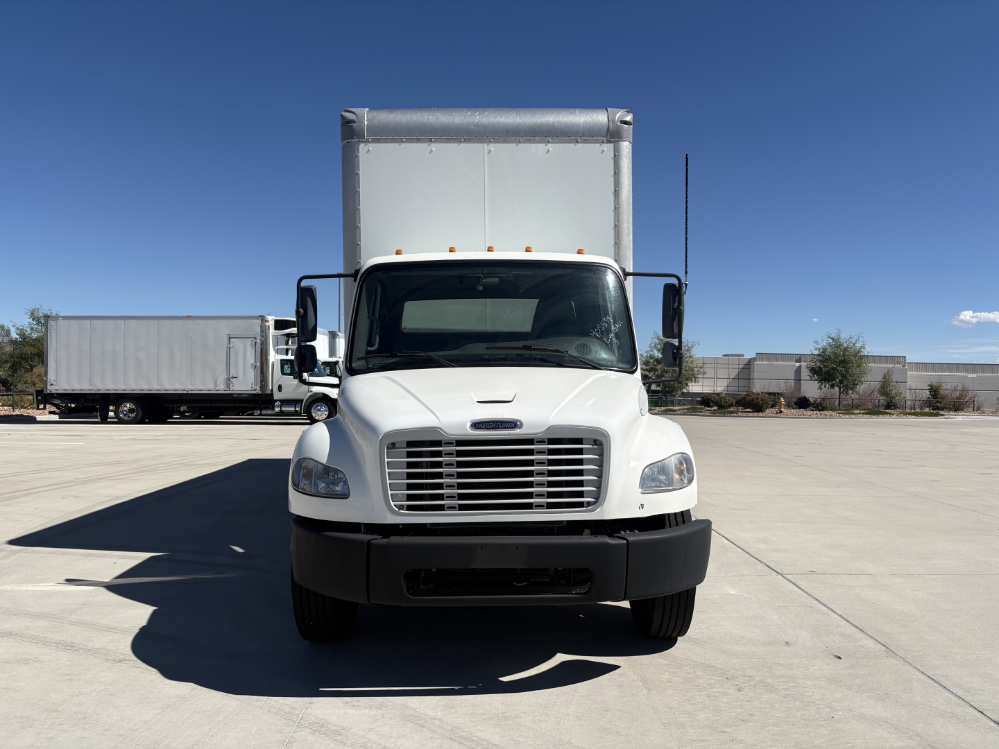 Freightliner M2-106 box truck front view showing grille and cab, with box trailer in background.