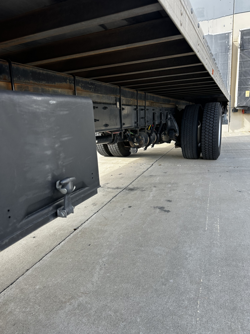 Freightliner M2-106 undercarriage showing dual rear wheels and trailer frame, side view of truck chassis and storage area, concrete ground.