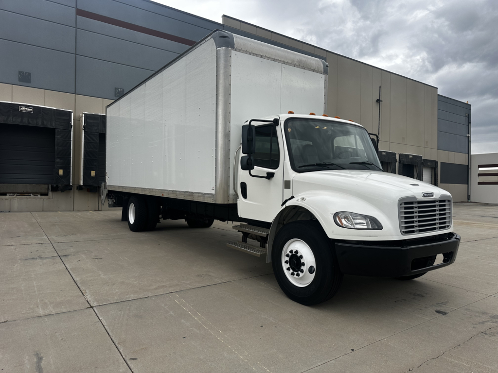 Freightliner M2-106 box truck in a dealership lot, side view showing white cab and large rectangular cargo box.