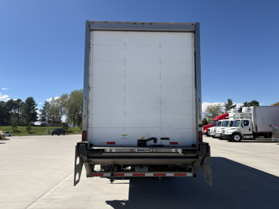 Freightliner M2-106 box truck rear view showing closed cargo door, rear bumper, and taillights; dealership lot in background.
