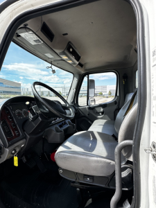Cab interior of a Freightliner M2-106 showing driver seat, steering wheel with dashboard, overhead compartments, and side mirror view.