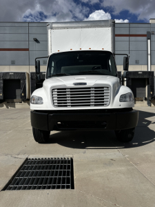 Freightliner M2-106 front view showing large chrome grille, white cab, black bumper, and side mirrors.