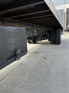 Freightliner M2-106 undercarriage showing dual rear wheels and trailer frame, side view of truck chassis and storage area, concrete ground.