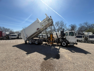 Truck side view of a 2018 Autocar XPeditor dump truck with bed raised, white cab, in a lot with other trucks under blue sky.