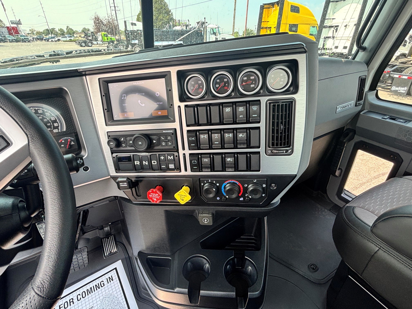 Truck dashboard interior showing instrument cluster, switches, and controls in Mack PI64T cab; steering wheel left, gear selector area, and cup holders visible.
