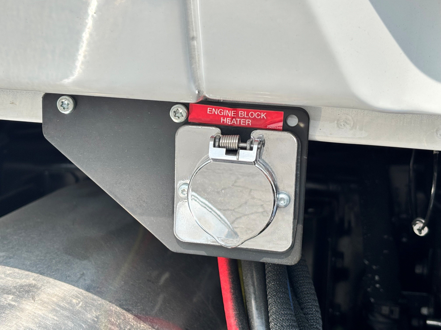 Engine block heater with red label reading 'ENGINE BLOCK HEATER' on a Mack PI64T cab and chassis showing a square metallic plug housing.