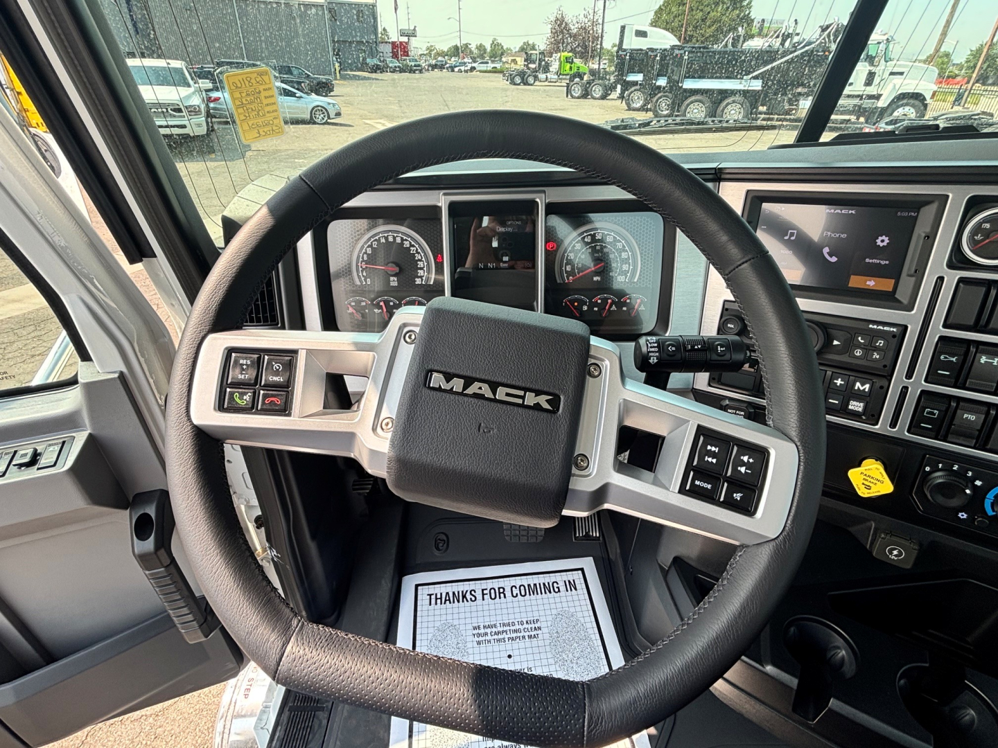Mack PI64T truck interior showing steering wheel with Mack logo, instrument cluster, and center dashboard. Accessories include controls on steering wheel and a screen on the right.