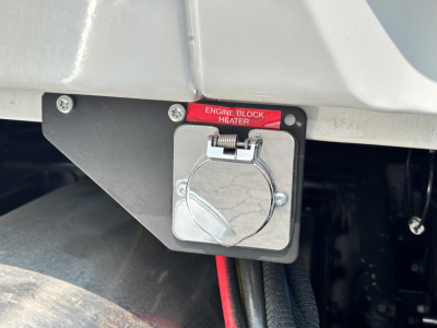 Engine block heater with red label reading 'ENGINE BLOCK HEATER' on a Mack PI64T cab and chassis showing a square metallic plug housing.