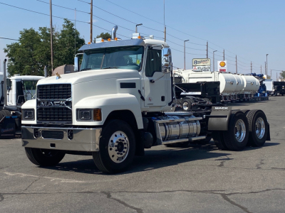 Front-left view of a white Mack PI64T truck with a long wheelbase, showing Mack grille and cab. Visible text includes MA CK on grille, wheels, and a tanker trailer in background.