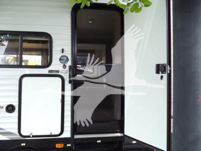 Travel trailer entry door open showing interior doorway and exterior storage hatch, with stickers near door frame.
