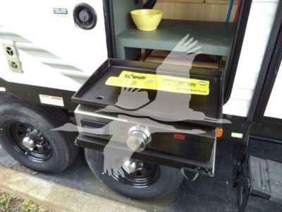 Trailer hitch area of a travel trailer with a yellow label on a fold-out shelf and a round hitch ball, side of the trailer with white exterior and black wheel.
