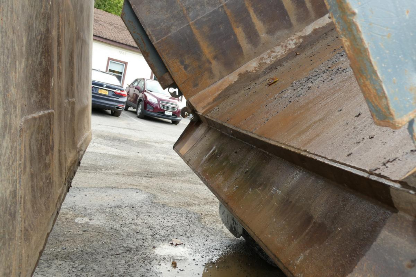 Dump truck bed side view with rusted steel blade and tip, gravel ground, vehicles in background.