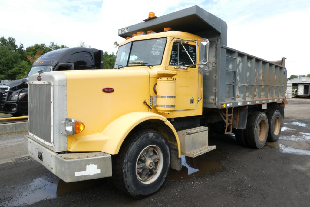 Peterbilt dump truck in use showing cab and dumping dump bed, yellow cab with chrome grille and large front bumper, VIN 1XPALB0X3JN264708 visible on windshield area.