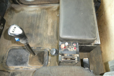 Truck interior: gear shifter with H pattern and labels, center console switches, and a rectangular black box with red/white buttons; visible floor mat and seating edge.