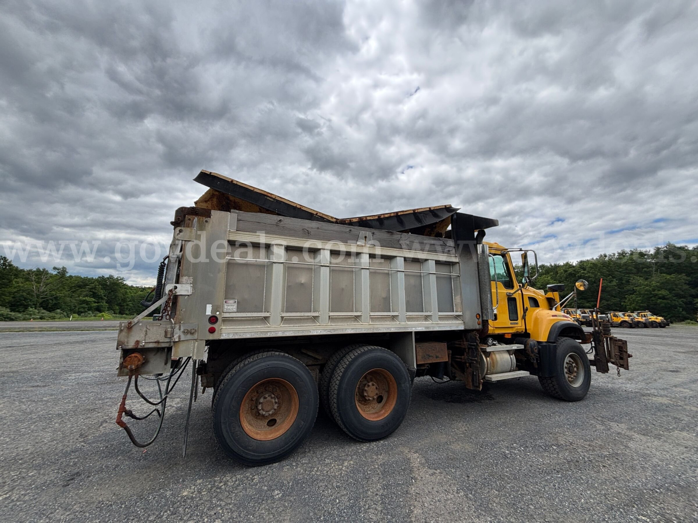 Dump truck with yellow cab and gray dump bed, three axles, end dump, side view showing rear wheels and hydraulic bed, daytime overcast sky.