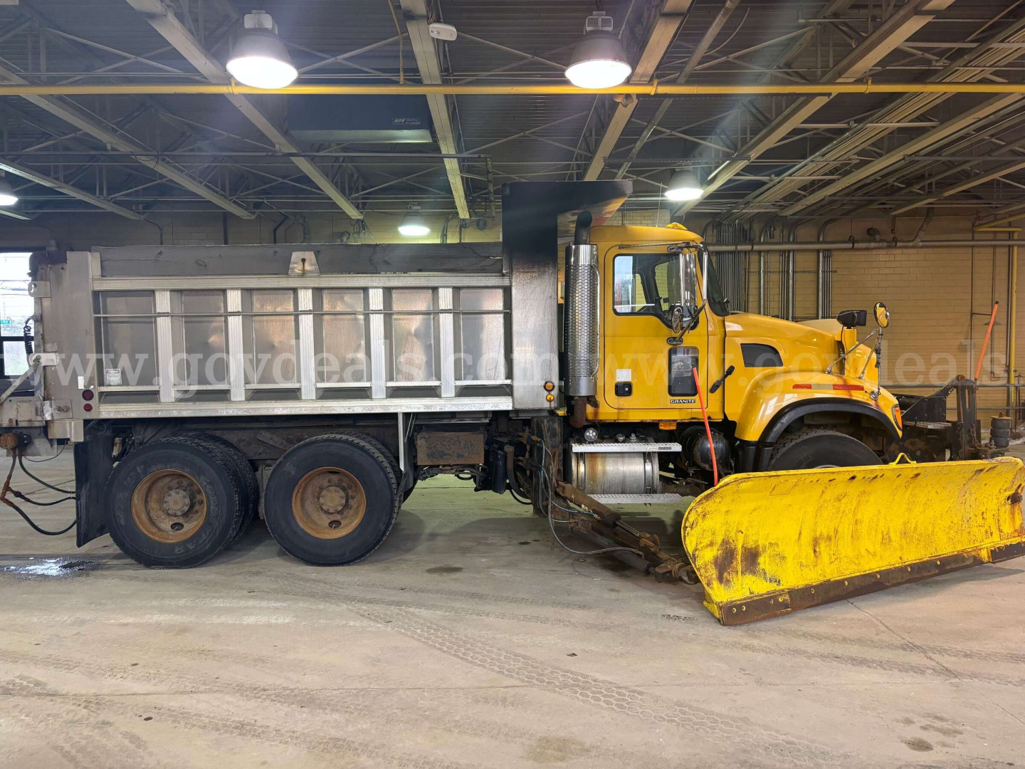 Mack CV713 dump truck in an indoor shop with a large yellow snow plow blade attached to the front; side view shows the cab and dual rear axles, metal dump bed.