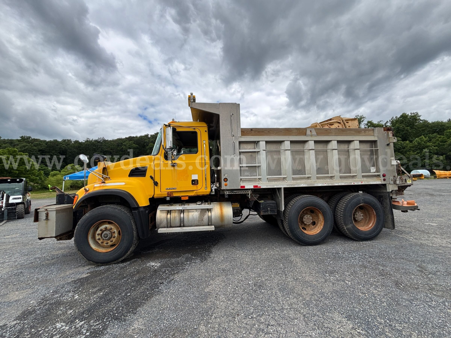 A yellow Mack CV713 dump truck with a gray dump bed parked on a gravel lot. Side view shows the cab, chrome fuel tanks, and dual rear axles. The truck has a front chrome bumper and a large exhaust. The sky is cloudy.