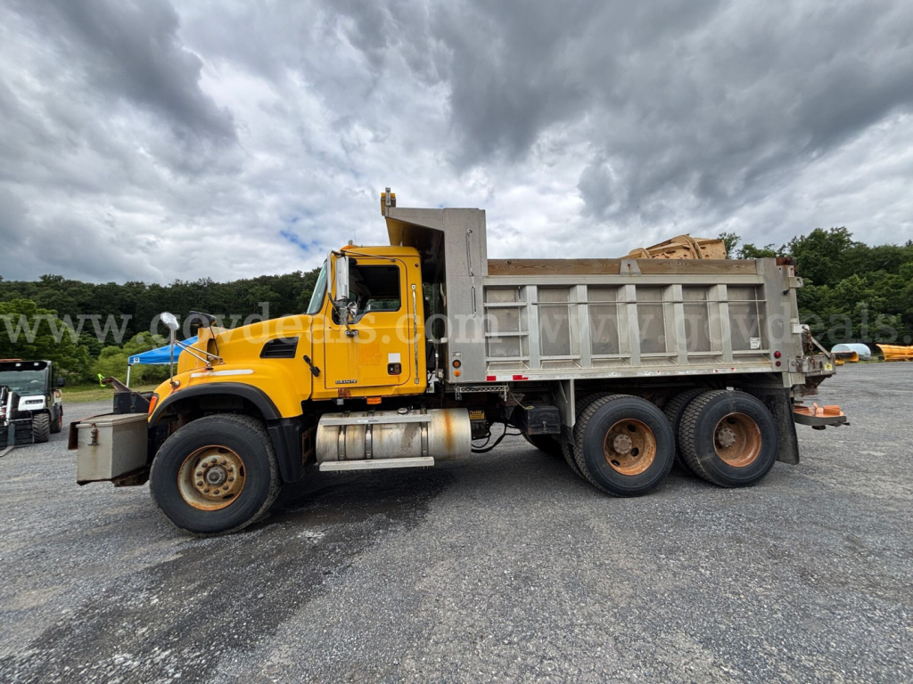 A yellow Mack CV713 dump truck with a gray dump bed parked on a gravel lot. Side view shows the cab, chrome fuel tanks, and dual rear axles. The truck has a front chrome bumper and a large exhaust. The sky is cloudy.