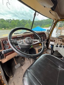 Cab view of 2006 Mack CV713 dump truck showing steering wheel, dashboard with wood trim, accelerator/brake pedals, and gear shifter.