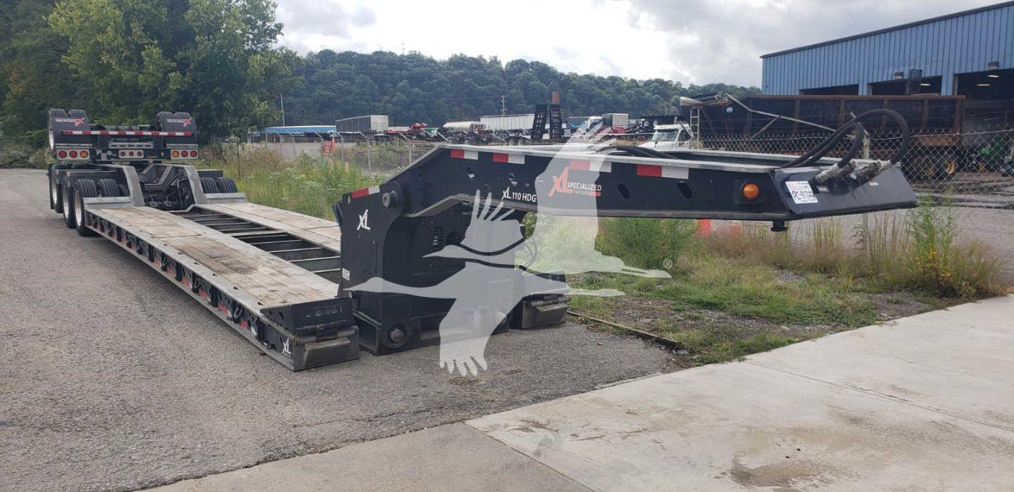 Lowboy trailer in yard showing XL Specialized XL110Hdgs15 with long black deck and hydraulic gooseneck, trailer and counterweight visible