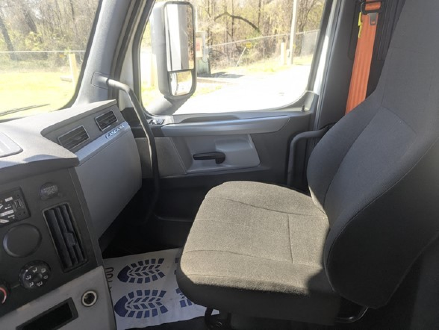 Driver seat and dashboard of a Freightliner truck cabin with gray interior; visible are steering column, dashboard controls, and a black fabric seat.