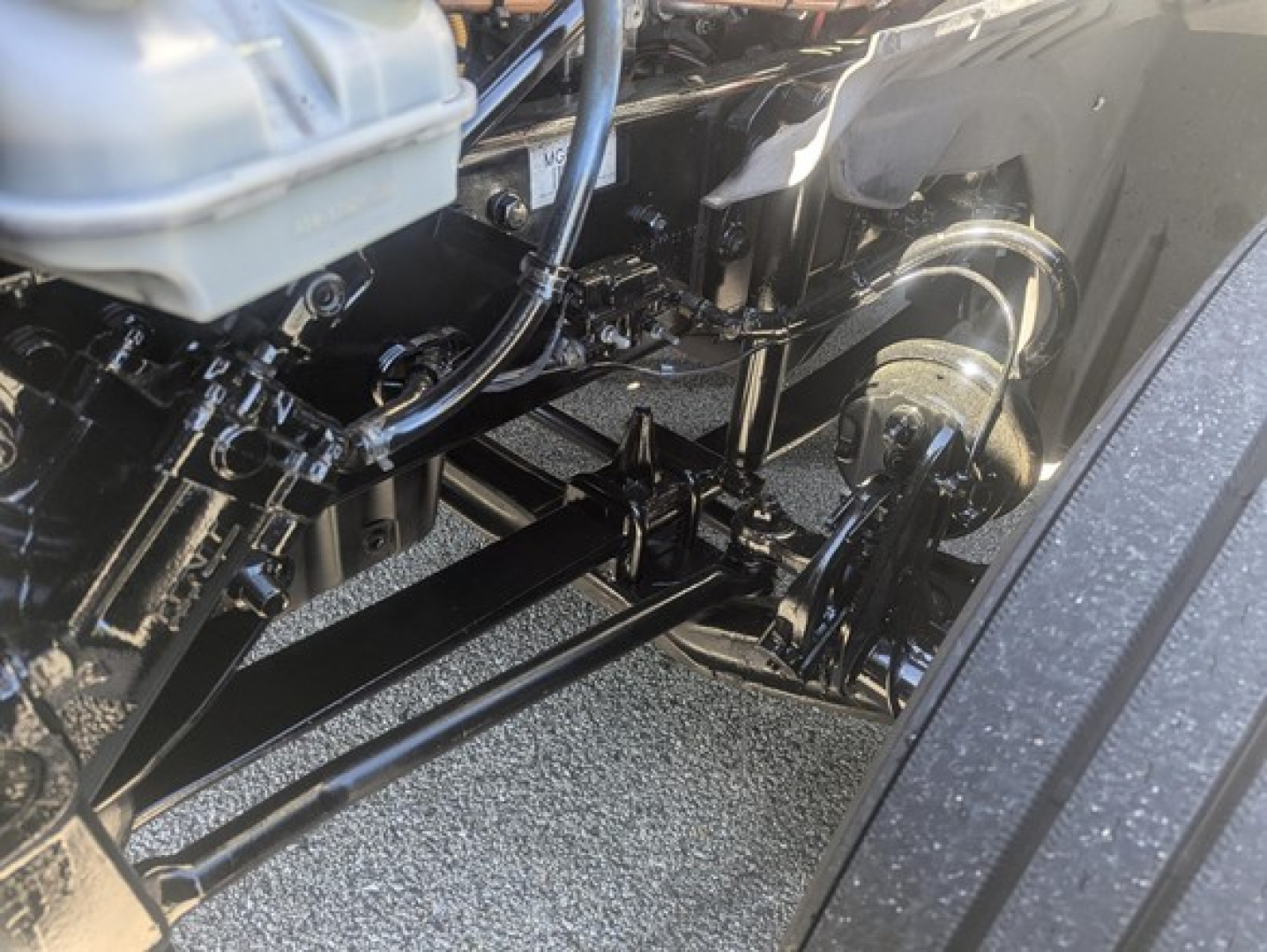 Truck front suspension and steering linkage in an engine bay view showing metal rods and hydraulic lines.