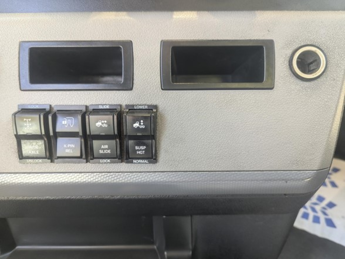 Dashboard panel of a Freightliner Cascadia showing various switches labeled AXL, PNL, REL, AIR SUSP LOCK, SUSP HGT, etc. Two rectangular slots and a circular keyhole on the right.