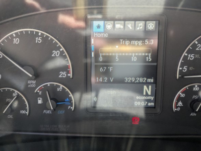 Dashboard of a 2021 Freightliner Cascadia 126 showing instrument gauges and a digital display. Text visible includes Home, Trip mpg: 5.3, 67 °F, 14.2 V, 329,282 mi, N gear, mph scale, and icons on the screen.