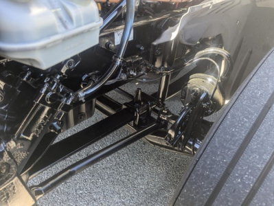 Truck front suspension and steering linkage in an engine bay view showing metal rods and hydraulic lines.